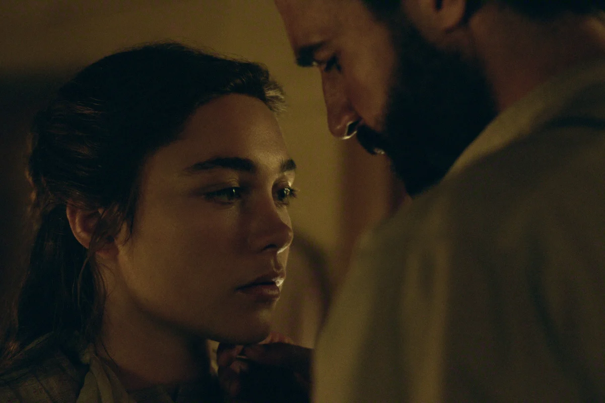 "A young woman and a bearded man stand closely in a dimly lit room, sharing an intense and intimate moment. The scene has a soft, warm glow and a serious mood, suggesting a private, emotional exchange between the two."